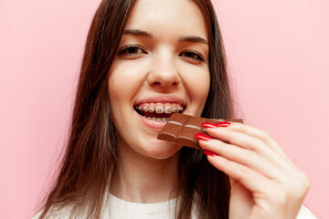 cheerful woman with braces eats chocolate and smiles on pink isolated background, young girl bites...