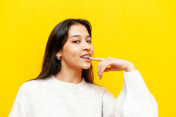 young pensive asian girl with braces flirts and thinks sexually over yellow isolated background, excited korean girl dreams and imagines and points finger at lip - obrazy, fototapety, plakaty