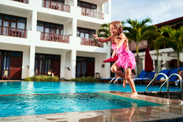 Excited girl in pink swimsuit jumps off poolside into clear blue water. Child enjoys summer swim,...