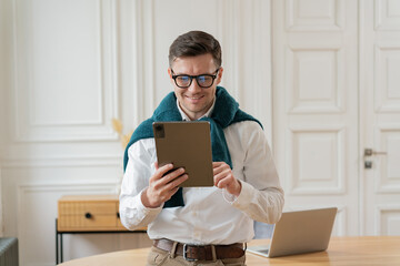 A poised man clad in business casual attire reviews a tablet with a smile, standing in a...