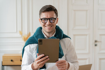 A cheerful man with glasses and a teal scarf engaging with a tablet in an elegant room suggests a...