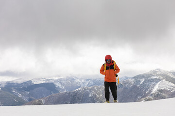 A man in an orange jacket stands on a snowy mountain. The sky is cloudy and the mountains are...