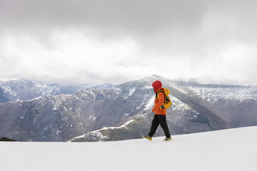 A person in an orange jacket is walking on a snowy mountain. The sky is cloudy and the mountains...
