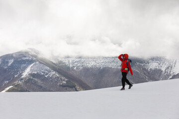 A person in a red jacket is walking on a snowy mountain. The sky is cloudy and the mountain is...