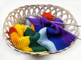 Colorful balls of wool for knitting, hand-knitted scarf in rainbow colors and knitting needles lie in a white wicker basket