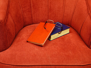 There are glasses, closed books and a burgundy decorative pillow in a red velour armchair 
