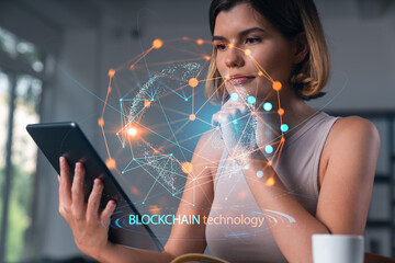 Smiling businesswoman in casual wear holding tablet device touching it at office workplace. Concept of distant work, business education, information technology.Blockchain hologram - Powered by Adobe