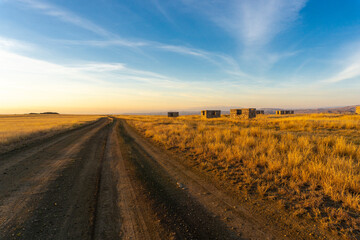 Evening view of an earthen road through the savannah and an abandoned village. Bright blue sky with clouds, orange grass and unfinished houses. Low mountains around. Georgia.