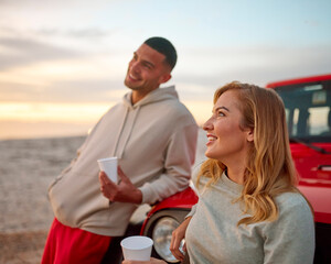 Obraz premium Young Couple Standing Chatting By Car With Hot Drink At Beach Watching Sunrise Together