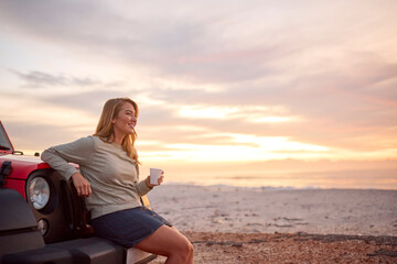 Fototapeta na wymiar Young Woman Standing By Car With Hot Drink At Beach Watching Sunrise 