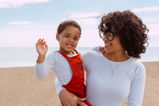 African mixed race attractive woman with curly hair and eyeglasses, holds her beautiful daughter at beach during spring. Baby says hello to camera with hand. Lifestyle concept.