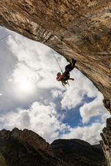 A person is hanging from a rock face with a rope. The sky is cloudy and the sun is shining