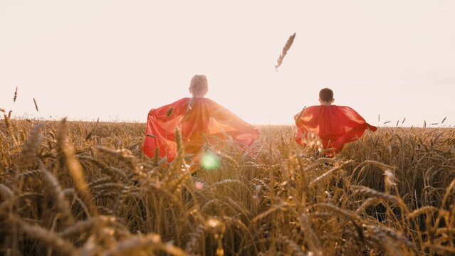 Happy kids in red superhero cloak running at sunlight dry wheat field back view. Joyful girl and boy playing flying hero fantasy imagination dreaming game playing enjoy freedom and childhood