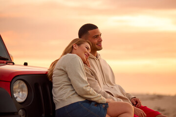 Loving Young Couple Sitting On Fender Of Car At Beach Watching Sunrise Together