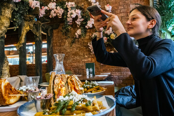 A young woman enjoys documenting her dining experience, taking a picture of a delicious sandwich in a charming cafe adorned with flowers