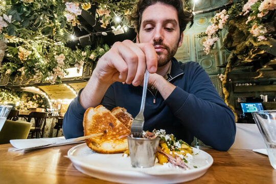 A man is engaged in enjoying a gourmet sandwich at a cafe, embellished with hanging garden decorations, offering a feast for the senses