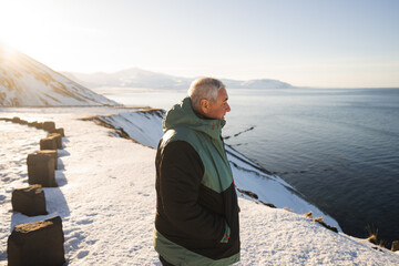 A pensive man wrapped in a warm jacket stands by the snowy seashore in Iceland, gazing at the calm ocean with mountains in the distance