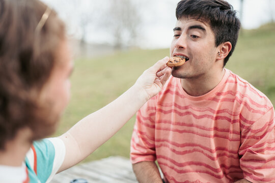 One man in a gay couple gently offers a cookie to his partner, a simple act of care and sharing between them in an outdoor setting