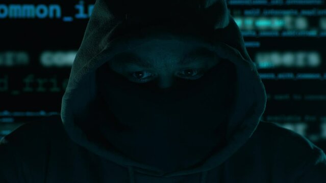 An anonymous hacker. Hooded figure with a masked face, program code in the background in a dark setting.