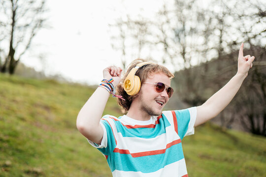 A cheerful man with a broad smile and yellow headphones raises his hand in joy while dancing in a serene outdoor setting