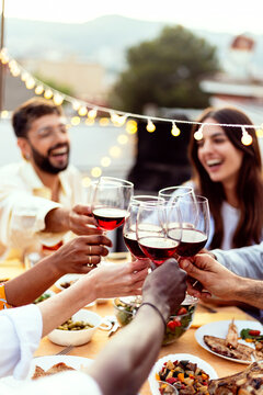 Group of young people toasting red wine in the evening at rooftop home party. Millennial diverse friends social gathering reunited on terrace table, having fun celebrating barbecue party at night.
