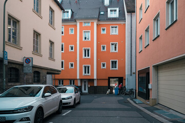 A white vehicle is parked in a tight alley between two orange buildings. Its automotive lighting...