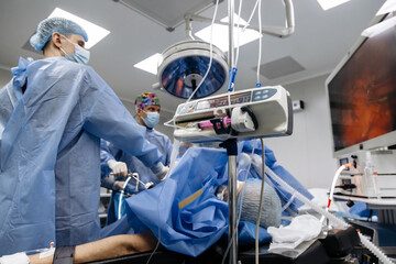 Experienced surgeons in medical gowns, caps, masks and gloves perform an operation in a modern clinic. Group of professional doctors using modern medical equipment in operating room.