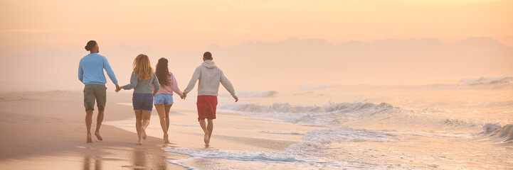 Rear View Of Couple With Friends In Casual Clothing On Vacation Running Along Beach Holding Hands