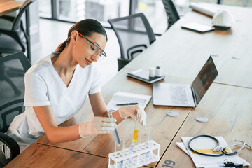 Scientist in glasses sitting by table with test tubes. Female doctor in white coat is indoors
