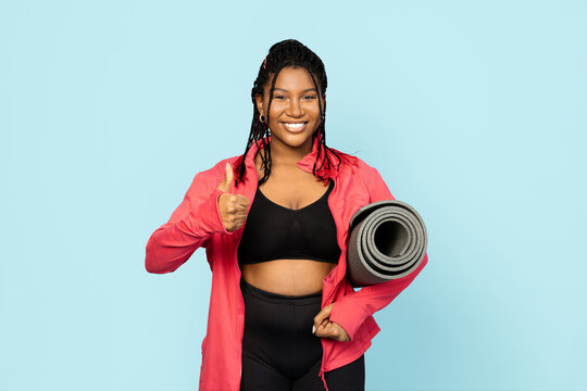 Fitness Woman Giving a Thumb Up. Confident woman in sportswear with a yoga mat against a blue background, signaling readiness for a workout.