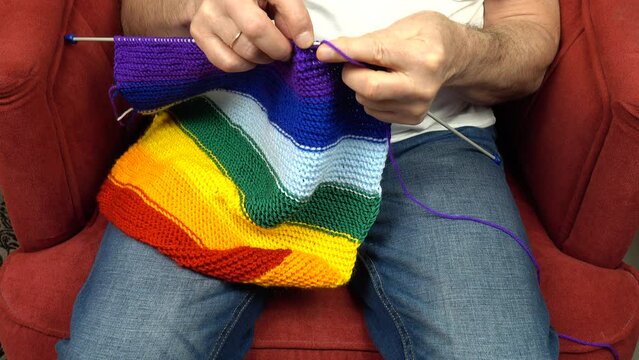 Person, sitting in an armchair, knits a scarf in rainbow colors on knitting needles 
