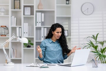 Worried Hispanic woman working in a home office setup reacts to bad news on her laptop, conveying...