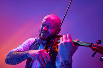 Bearded bald man, musician emotionally playing violin against gradient purple background in neon...