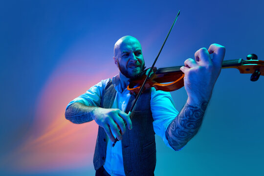 Deep intense melody. Artistic bald man, musician playing violin against blue background in neon with mixed light. Concept of music, talent show, performance, concert, festival, instruments