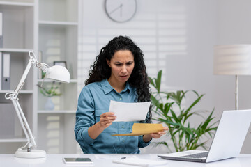 Concerned Latina businesswoman reviewing a document with bad news in a modern home office setting, expressing stress and frustration.