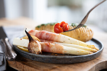 Fried white asparagus wrapped in bacon with herbed potatoes and tomatoes on black ceramic plate Short depth of field.