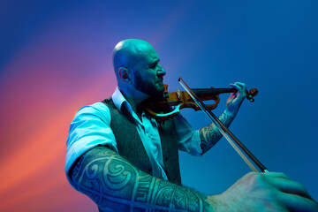 Deep feelings of melody. Young bald man playing violin against blue background in neon with mixed...