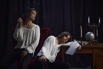 Portrait of man stands and smoking while another rests his head on desk in period-styled room with vintage furniture. Concept of comparisons of eras, renaissance, friendship, inspiration, afflatus.