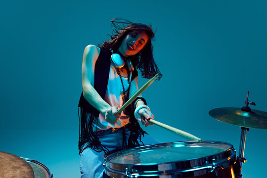 Energy burst. Young emotional, active woman, musician in stylish clothes playing drums against cyan background in neon light. Concept of music, talent show, performance, concert, festival, instruments - Powered by Adobe