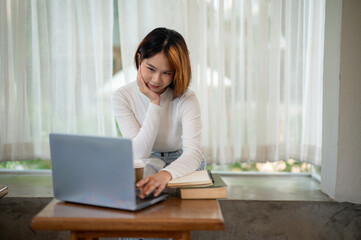 A cheerful young woman working on a laptop in a well-lit room with sheer curtains.
