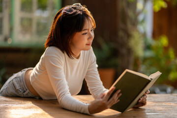 A young woman deeply absorbed in reading a book, bathed in warm sunlight at a rustic outdoor table,...
