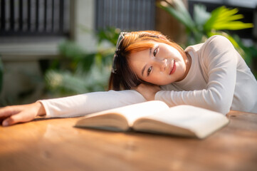 A dreamy young woman lies with her head on a wooden table, gazing thoughtfully beside an open book...