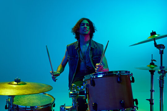 Artistic young boy in stylish clothes, musician playing drums against blue background in neon light. Rock and roll. Concept of music, talent show, performance, festival, instruments, youth culture