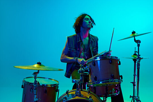 Young boy, musician in casual clothes playing drums, training against blue background in neon light. Concept of music, talent show, performance, concert, festival, instruments, youth culture