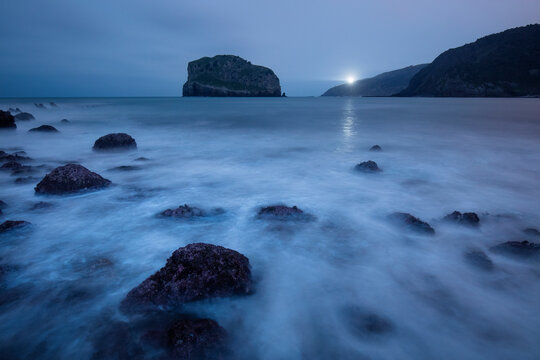 A solitary lighthouse beacon shines across the dusk-veiled waters near the rugged coast of Gaztelugatxe, Spain