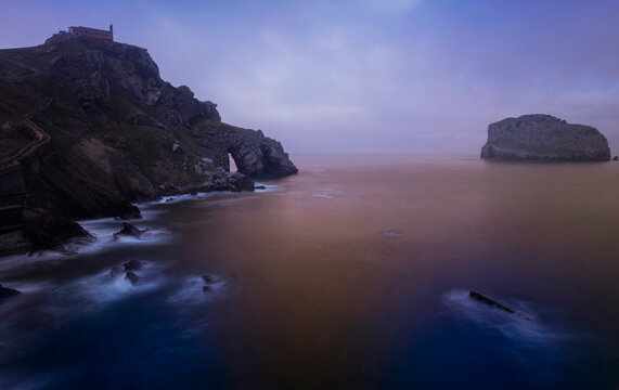 The hermitage of Gaztelugatxe perched atop a rugged cliff is enveloped in morning mist, with the sea whispering secrets to ancient rocks