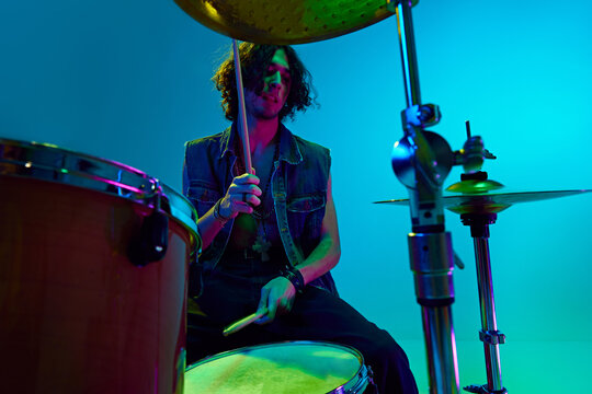 Artistic young boy in stylish clothes, musician playing drums against blue background in neon light. Rock and roll. Concept of music, talent show, performance, festival, instruments, youth culture