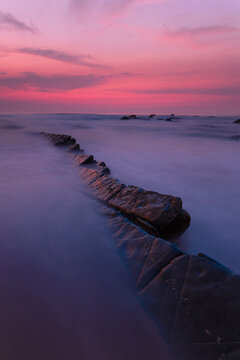 A natural rock pathway leads into the heart of a crimson sunset at Playa de Barrika, Bilbao, with the ocean waves softened into mist