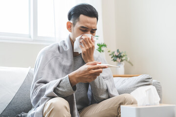 Sick, influenza asian young man have fever blowing nose, sneezing in tissue, holding thermometer...