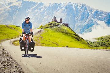 Male cyclist on touring bicycle adventure cycle visits Gergeti trinity church with mountains background. Cycling holidays and travel around caucasus mountain range - obrazy, fototapety, plakaty